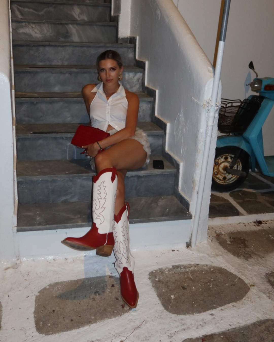 Georgie Young from Rallee Gummies in Mykonos, sitting on stone steps wearing red and white cowboy boots.