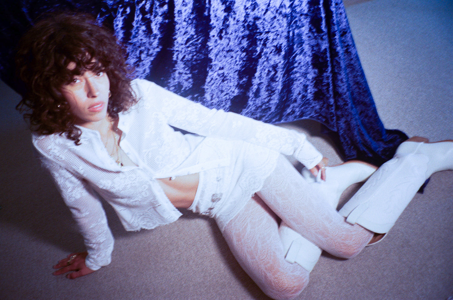 Person wearing a white lace skirt and top, and white cowboy boots, in front of a blue velvet sheet.