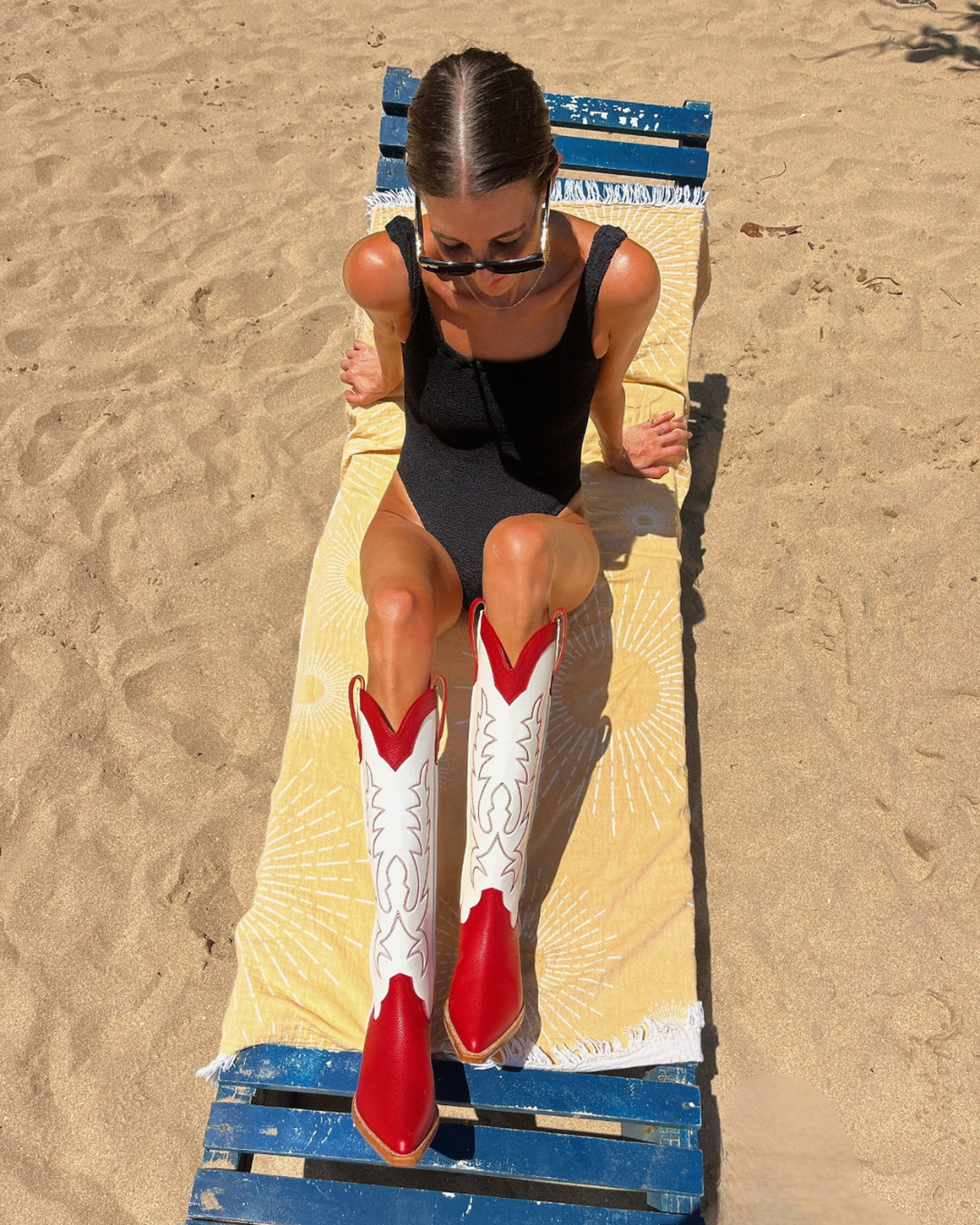 Woman in black swimsuit wearing red and white cowboy boots on a towel at the beach.