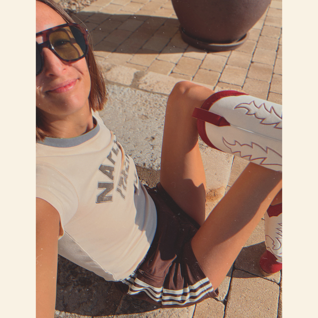 Woman sitting outdoors wearing sunglasses and a t-shirt with text, Adidas shorts and red and white cowboy boots.