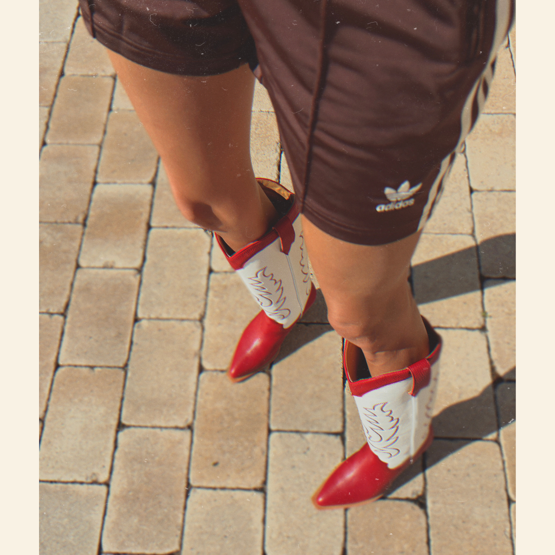 Person wearing red and white cowboy boots on a paved surface with brown Adidas shorts.