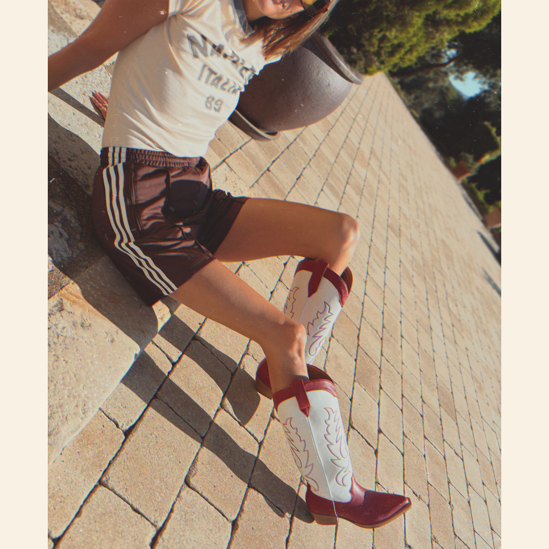 Person wearing a white t-shirt, brown Adidas shorts with white stripes, and red and white cowboy boots on a paved walkway.
