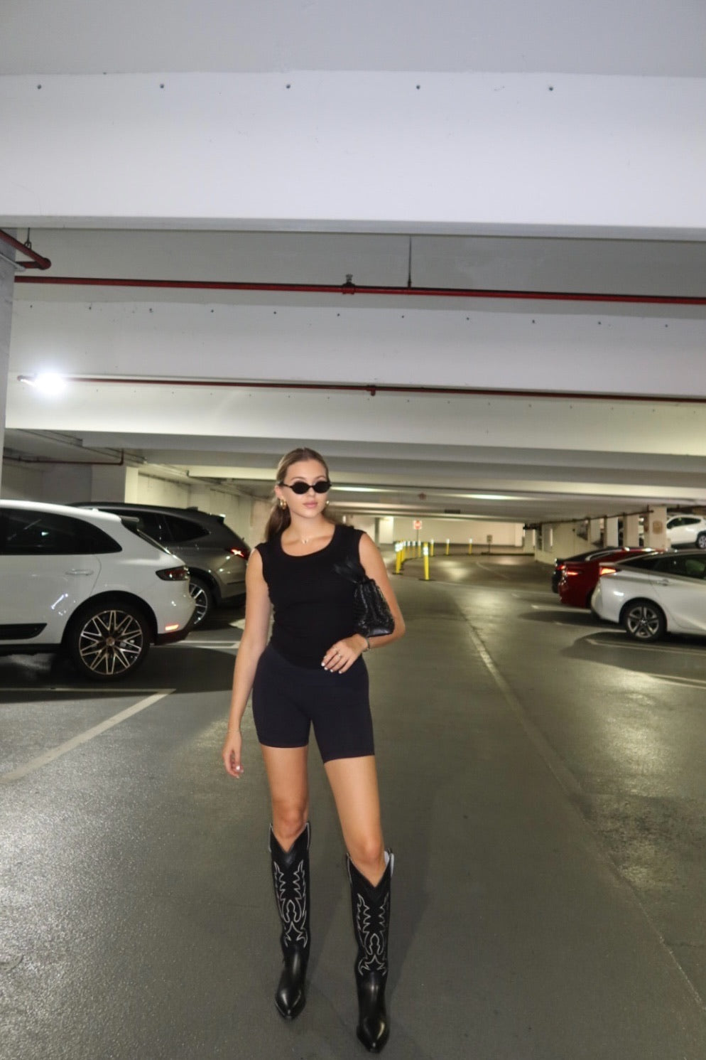 Woman in a black outfit with black cowboy boots standing in a parking garage.