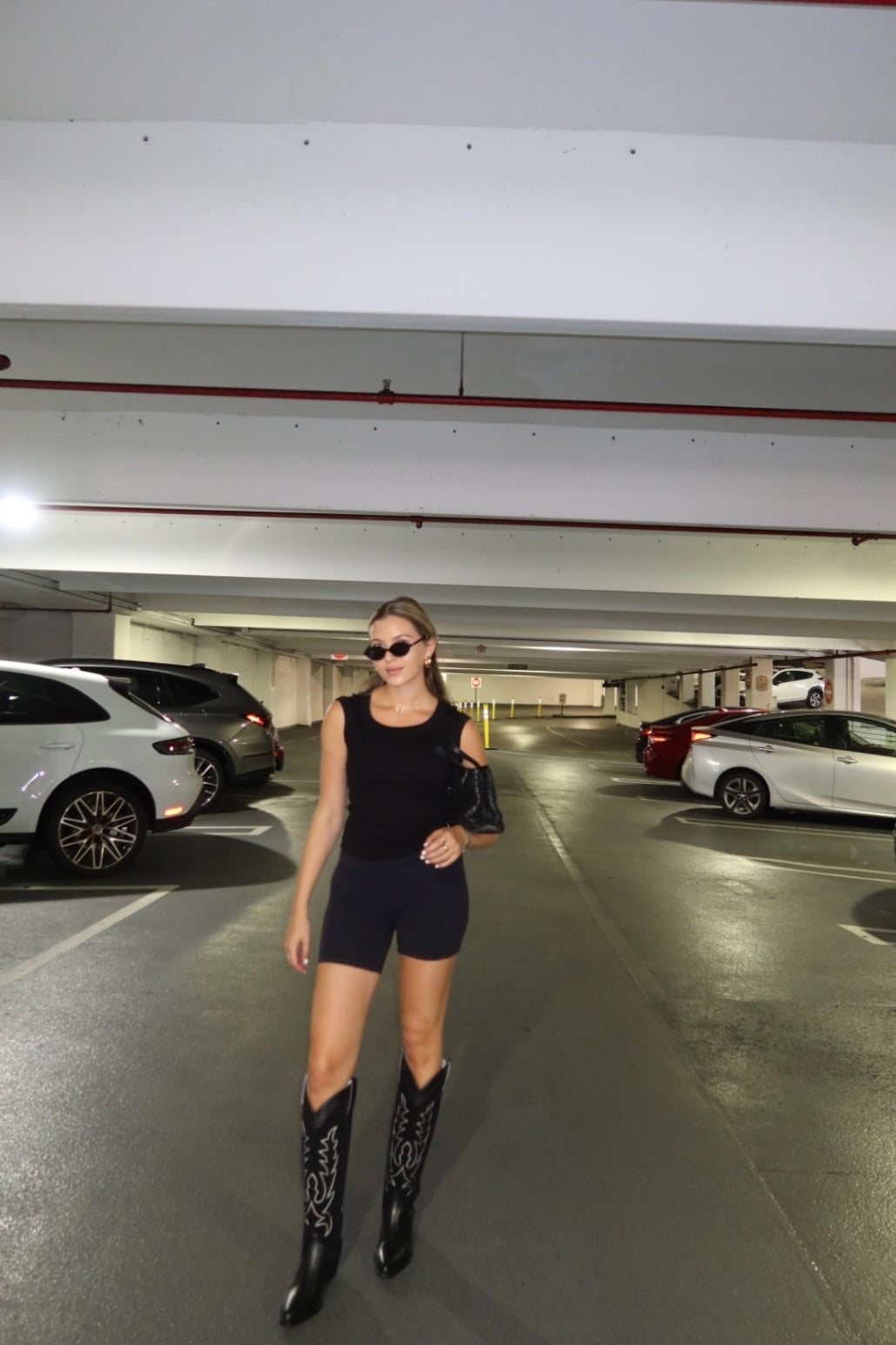 Woman in a black outfit with black cowboy boots standing in a parking garage.