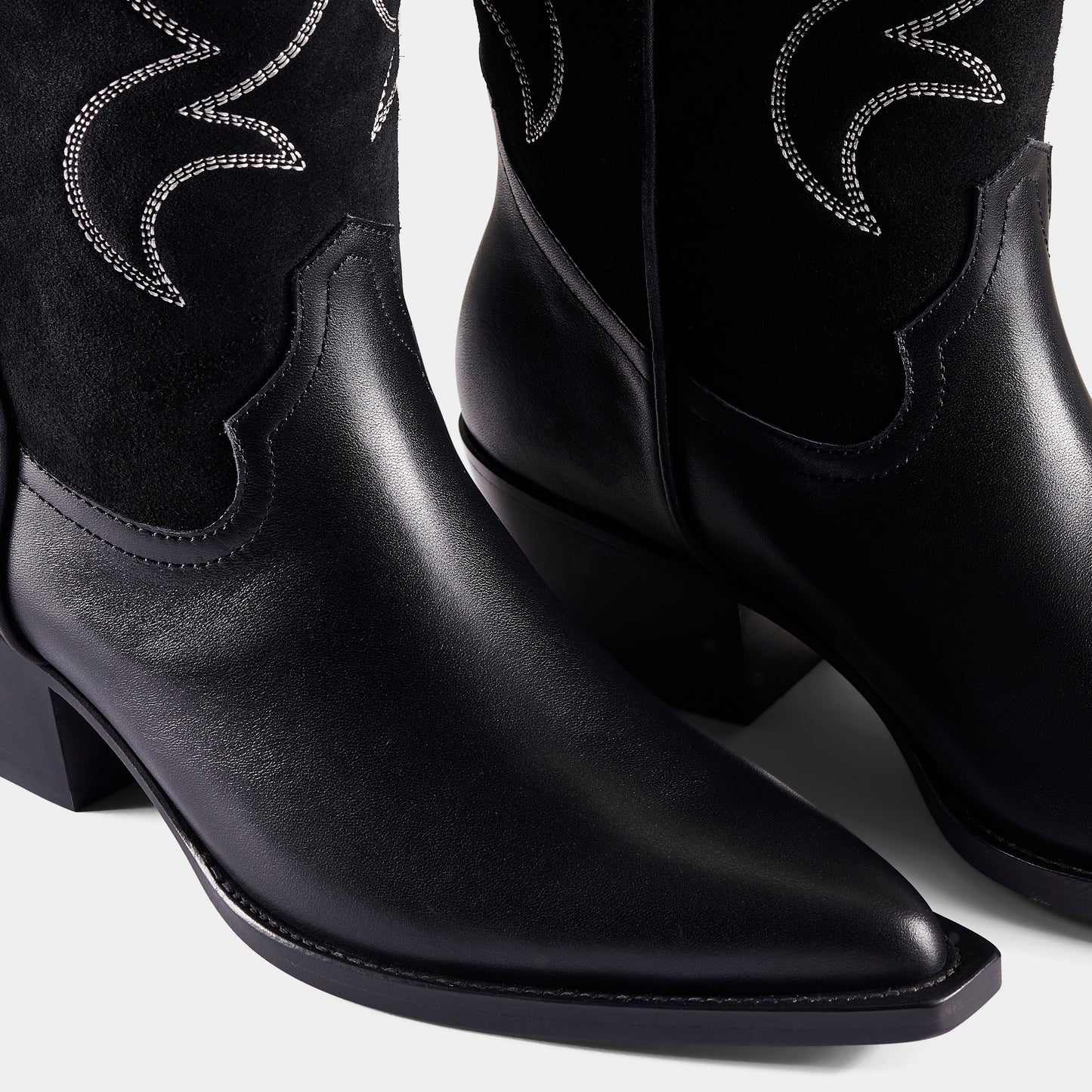 A black embroidered cowboy boot with a pointed toe, embroidered panels, and a low heel - upclose.