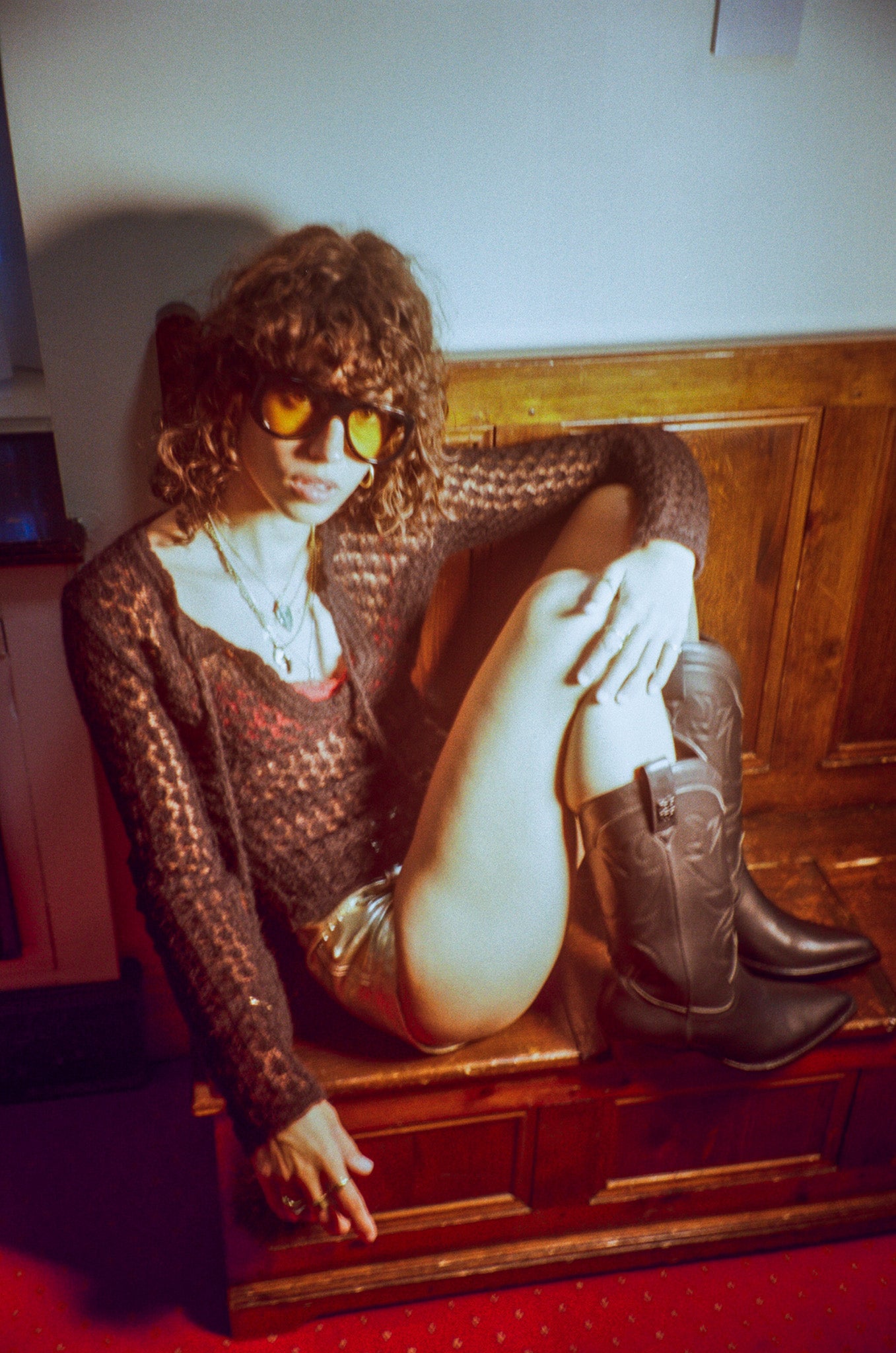 Person sitting on a wooden bench wearing sunglasses and black cowboy boots in a room with a wooden wall.