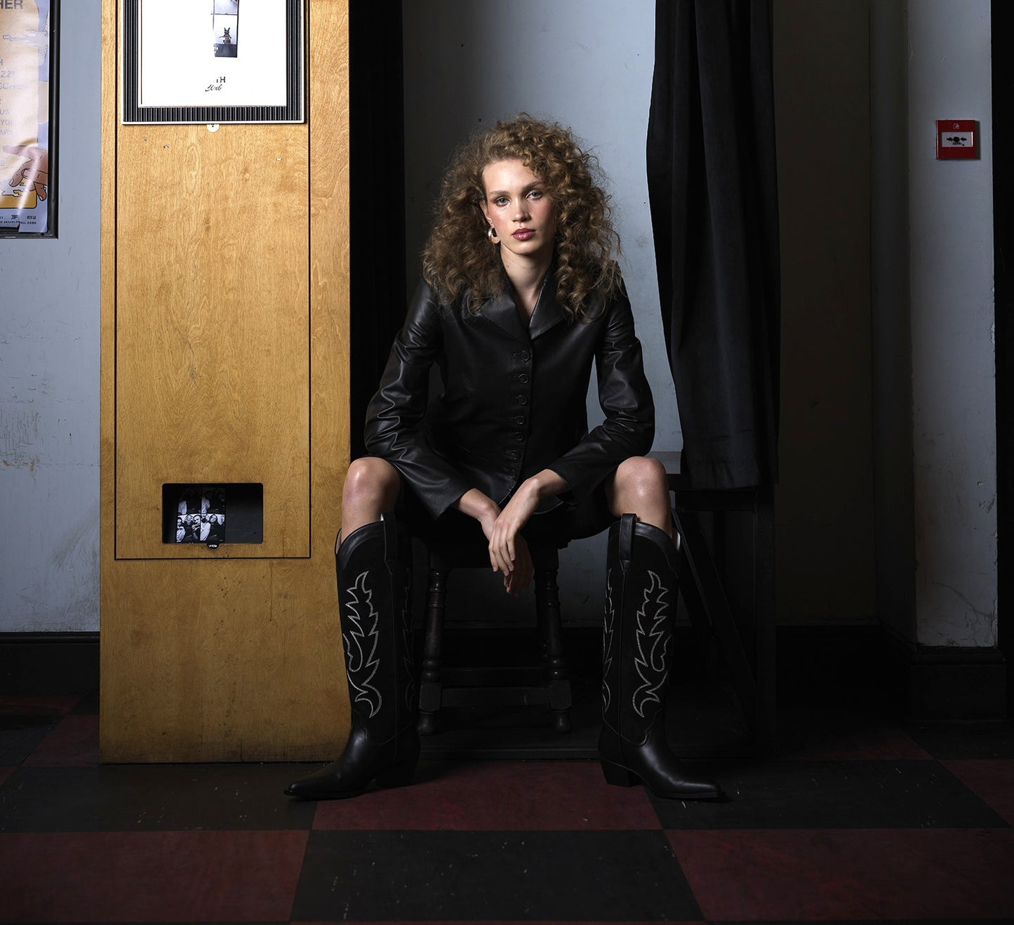Woman in black leather outfit and boots sitting in a dimly lit room.