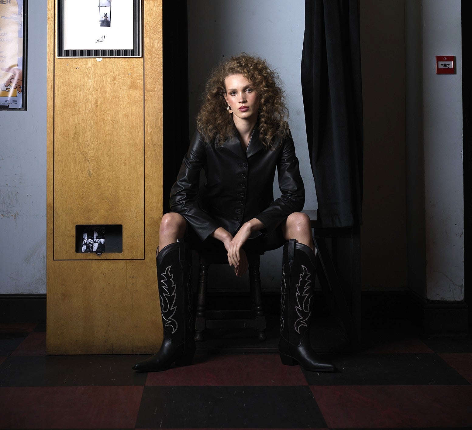 Woman in black leather outfit and boots sitting in a dimly lit room.