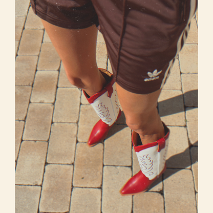 Person wearing red and white cowboy boots on a paved surface with brown Adidas shorts.