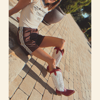 Person wearing a white t-shirt, brown Adidas shorts with white stripes, and red and white cowboy boots on a paved walkway.