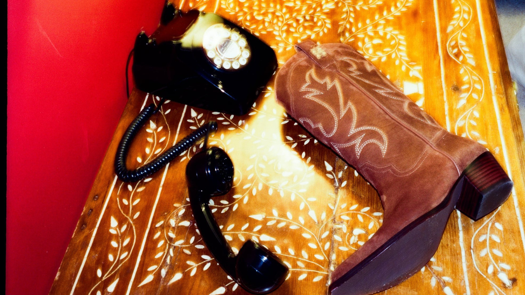 A brown suede cowboy boot with white western stitching lies on a carved wooden chest next to a black rotary telephone.