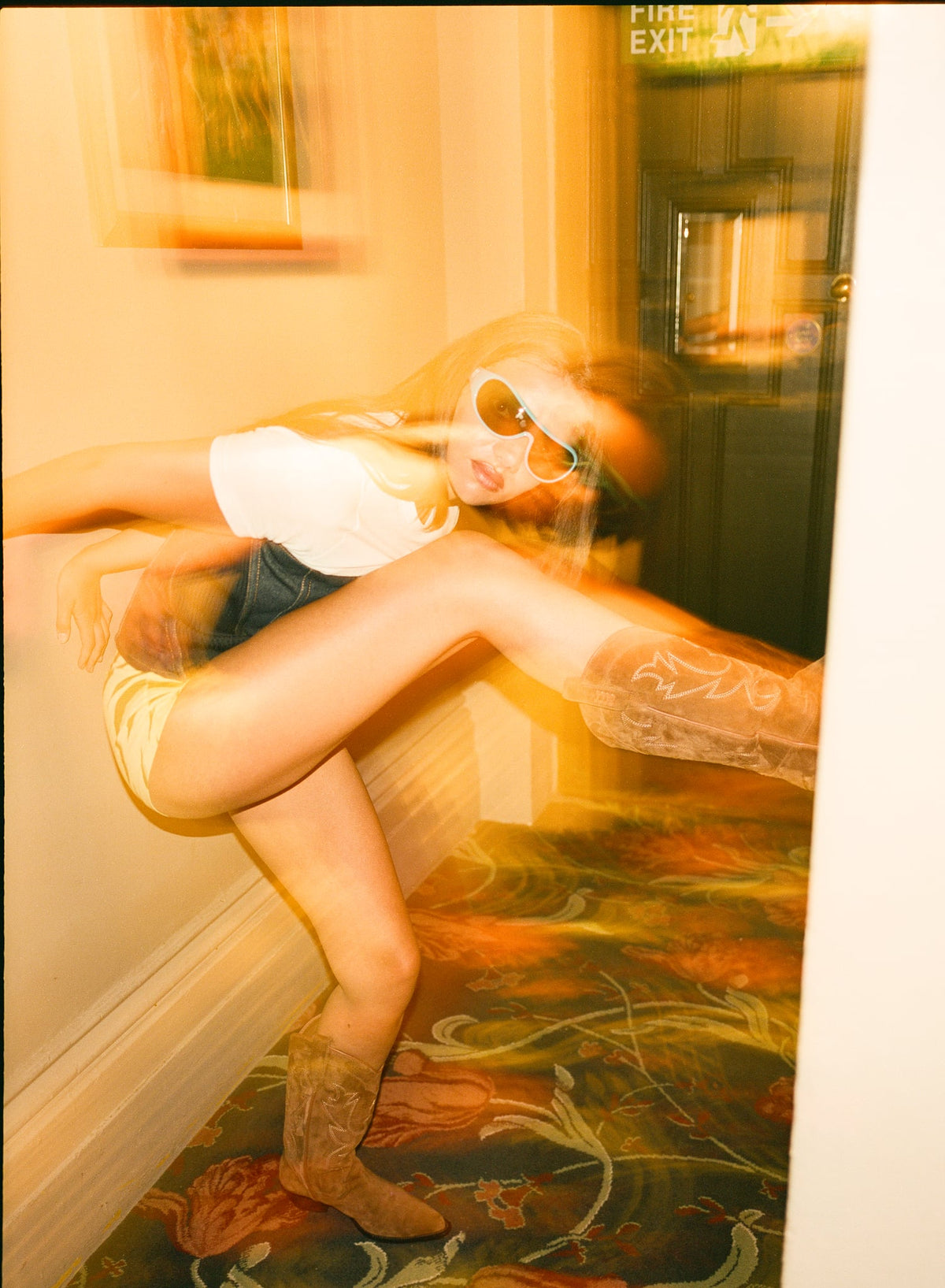 A woman in a white t-shirt, denim corset and animal print shorts, and cowboy boots poses dynamically in a hallway with a warm, yellow-orange filtered light and motion blur.