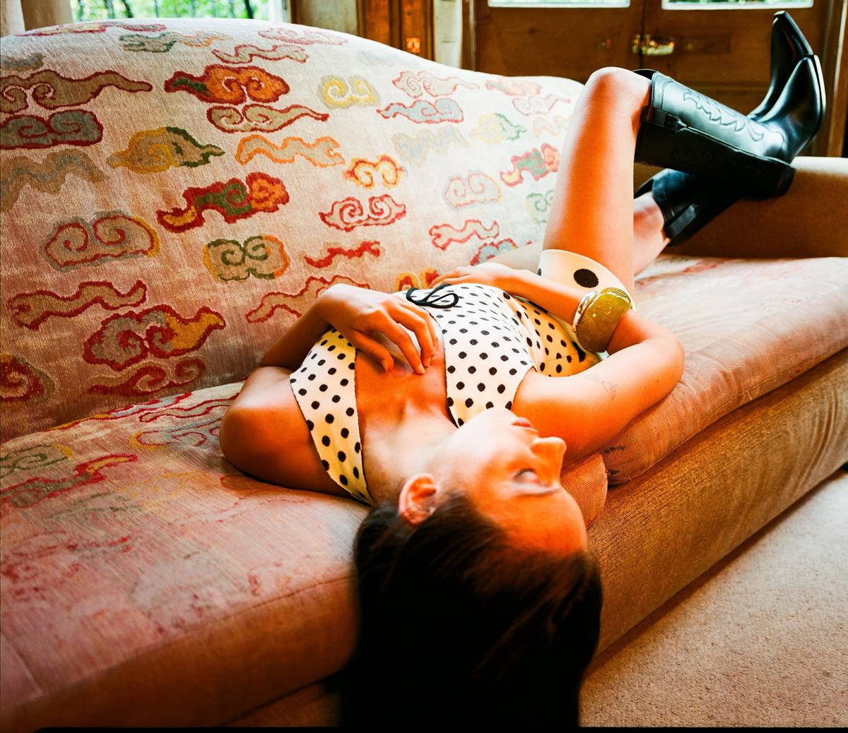 A woman reclining on a patterned sofa, wearing a polka dot top and  shorts, and black knee-high cowboy boots.