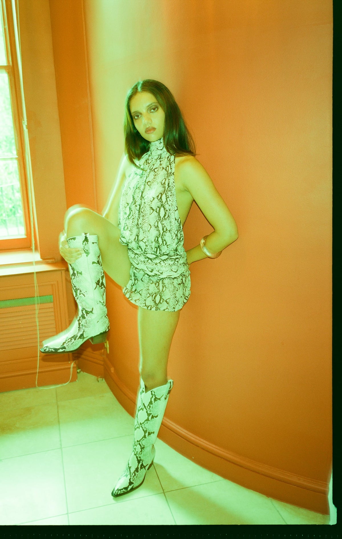 A woman posing in a matching snakeskin print sleeveless top and shorts set, paired with matching snakeskin print knee-high boots, set against a warm orange wall.