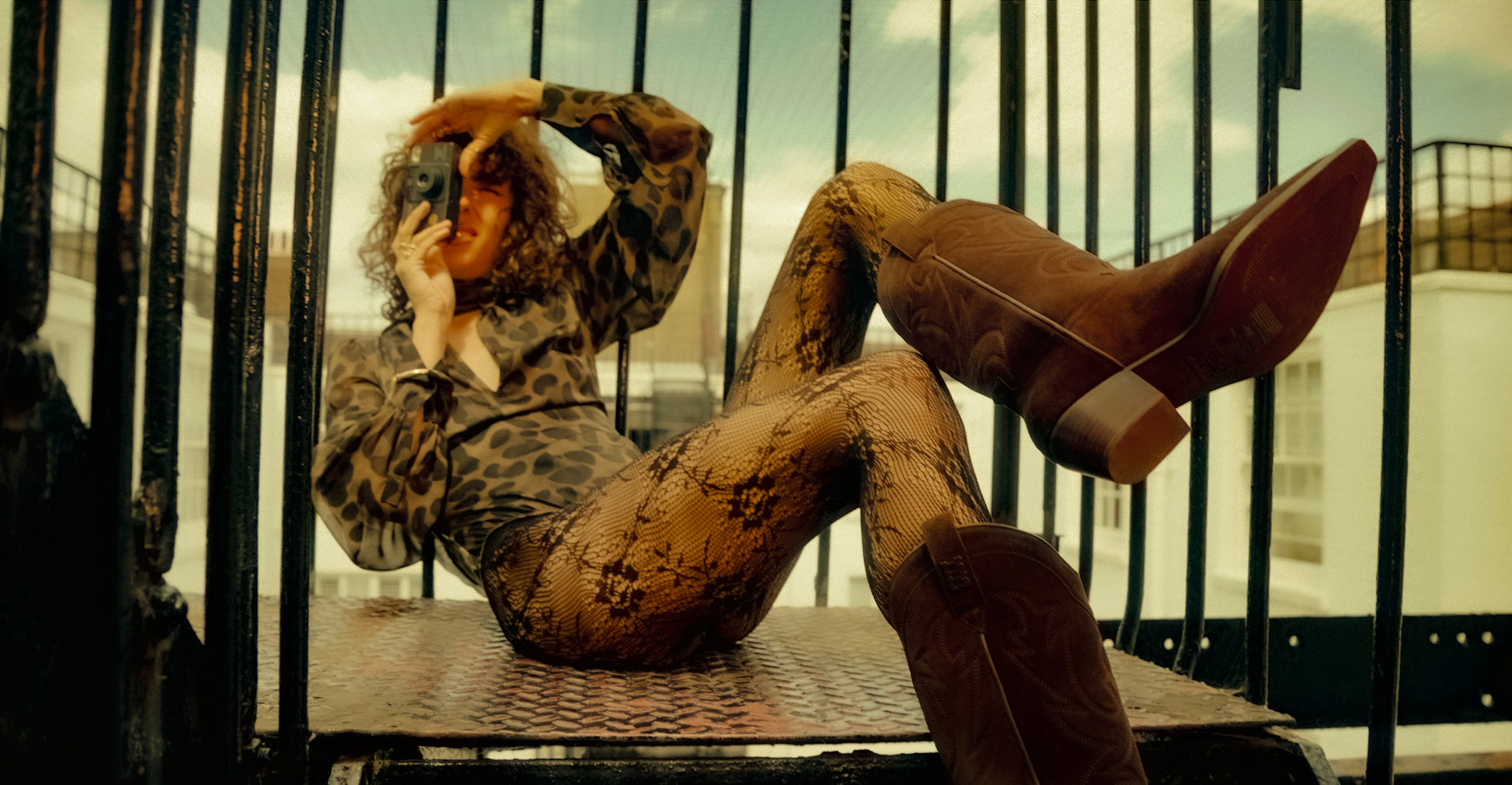 Person in Vespertine animal print body and brown cowboy boots sitting on a metal railing with a blurred background.