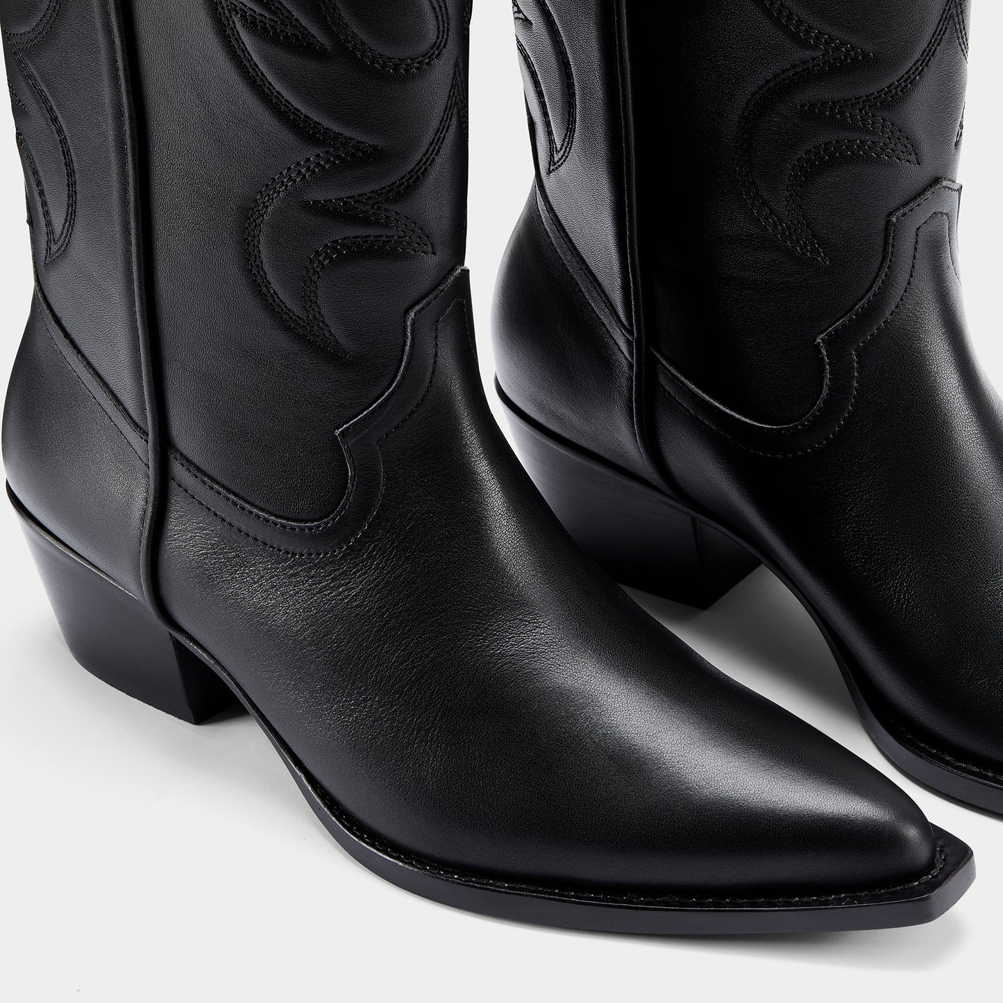 Black cowboy boot with embroidered design and metal branding on a light grey background - close up.