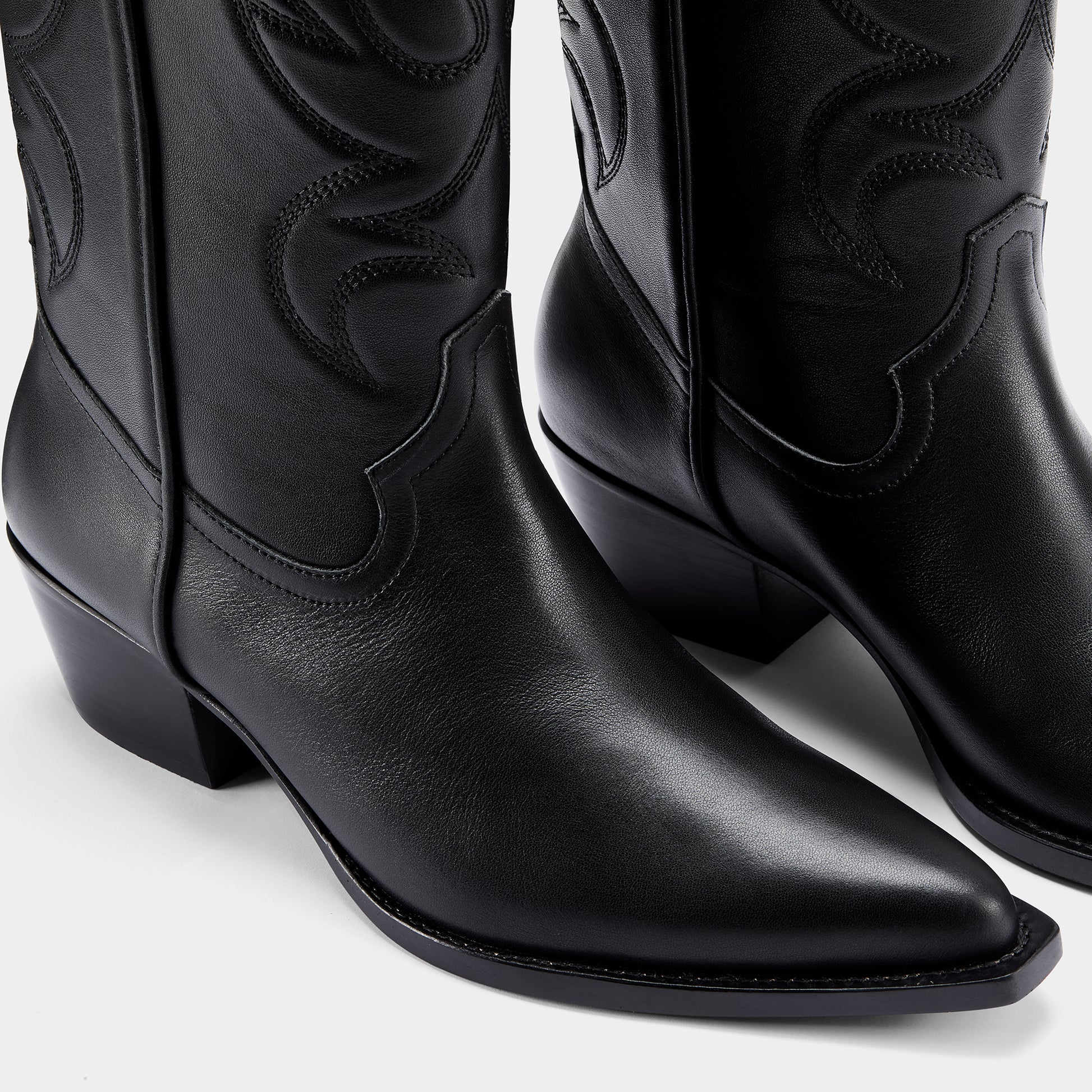 Black cowboy boot with embroidered design and metal branding on a light grey background - close up.