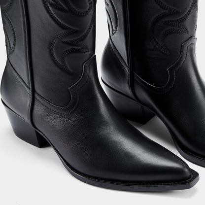 Black cowboy boot with embroidered design and metal branding on a light grey background - close up.