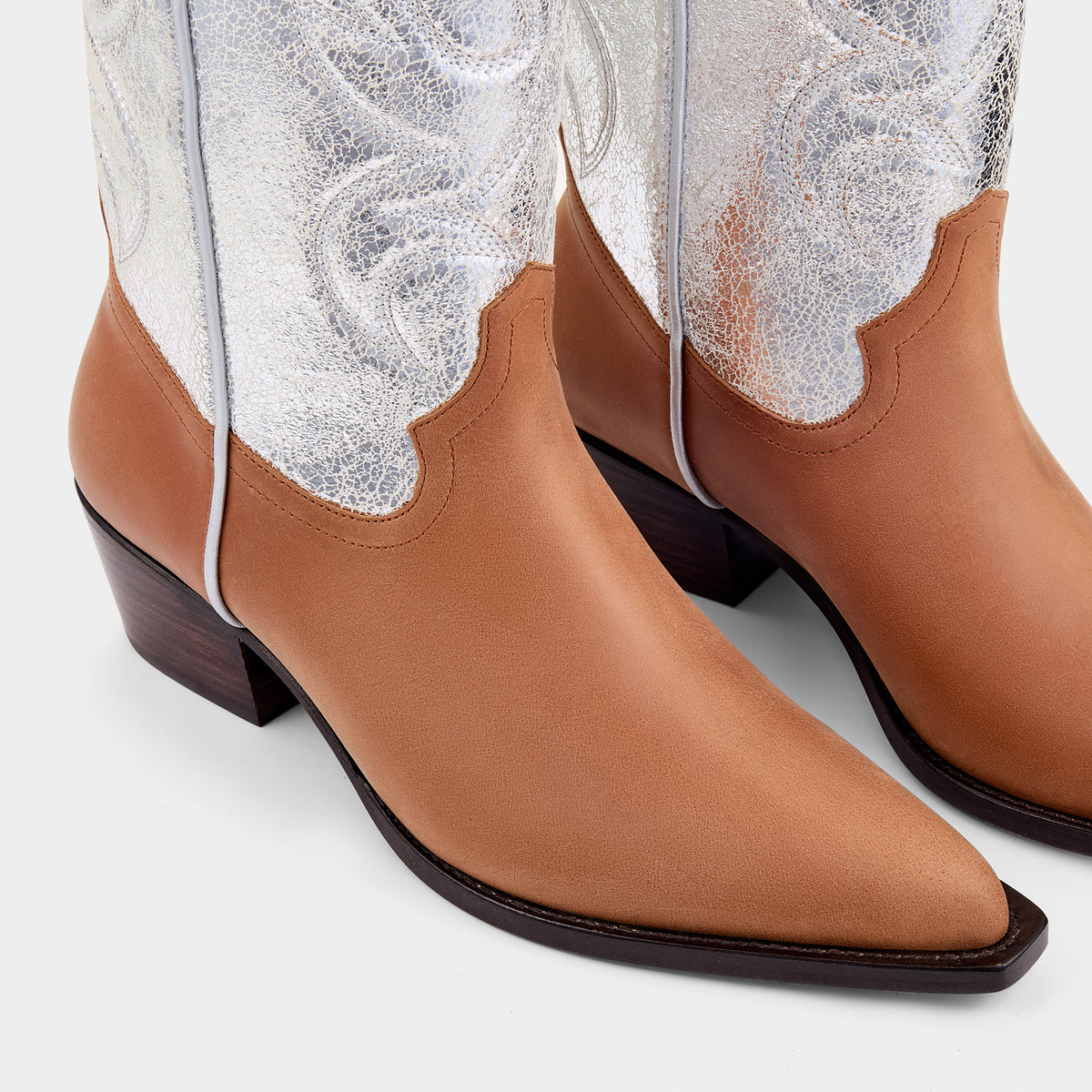 Tan and silver cowboy boot with cracked silver embroidered panels on a light grey background - close up