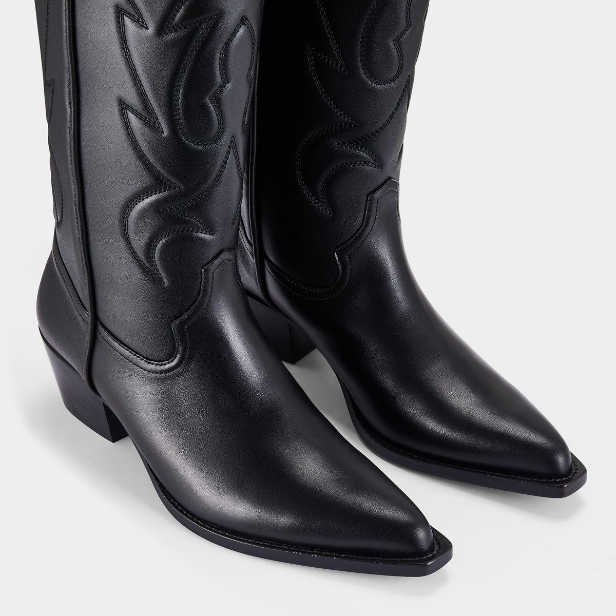 Black knee-high cowboy boot with a black embroidery design on a light grey background - close up