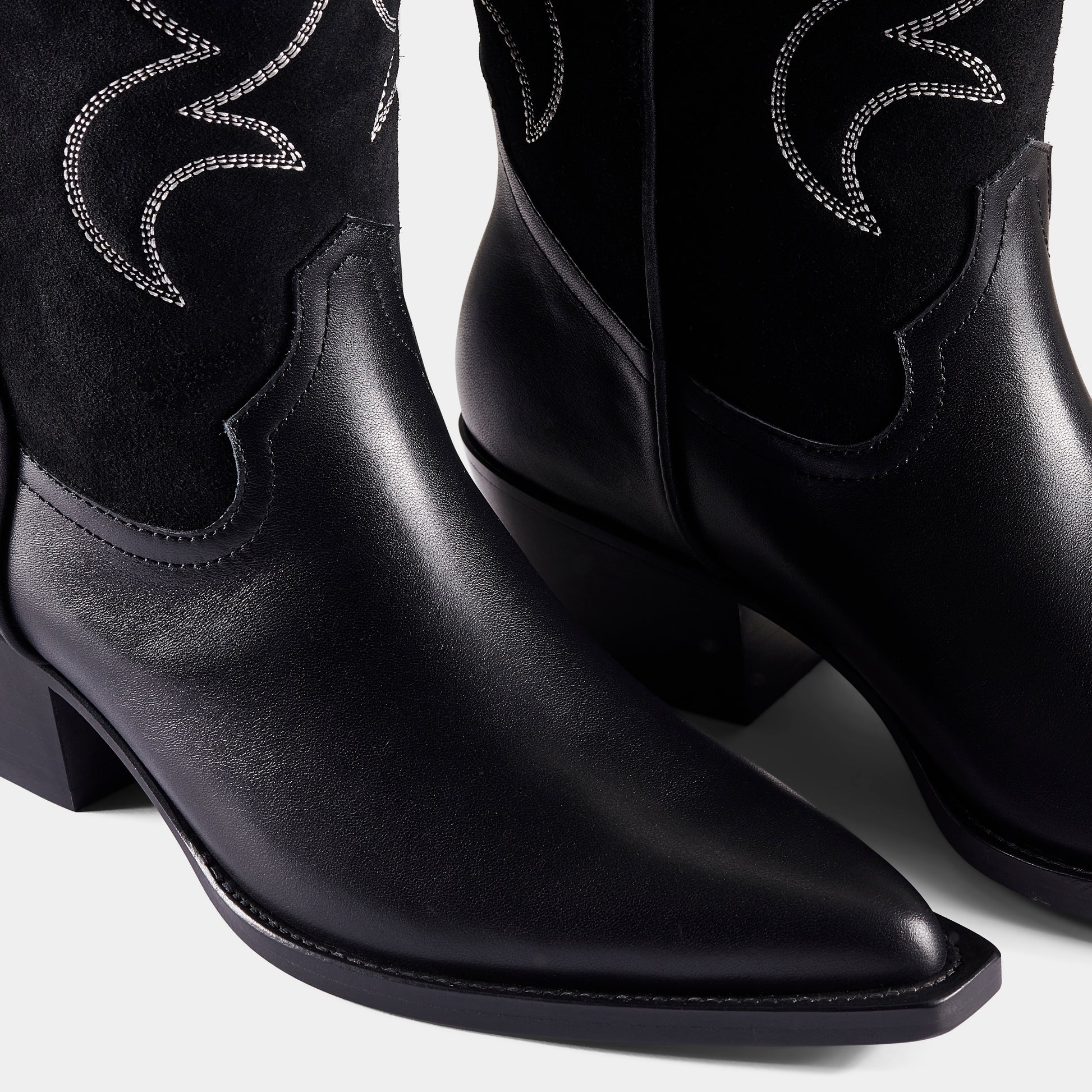 A black embroidered cowboy boot with a pointed toe, embroidered panels, and a low heel - upclose.